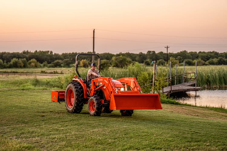 A man is driving a Kubota MX5400 in a field next to a pond